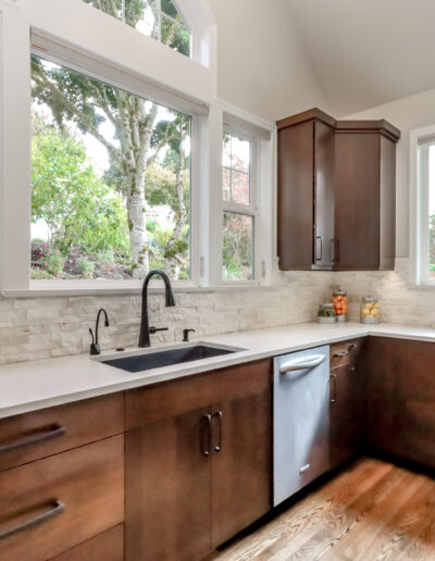 Modern kitchen with dark wood cabinets, light countertops, stainless steel appliances, and large windows overlooking a garden.
