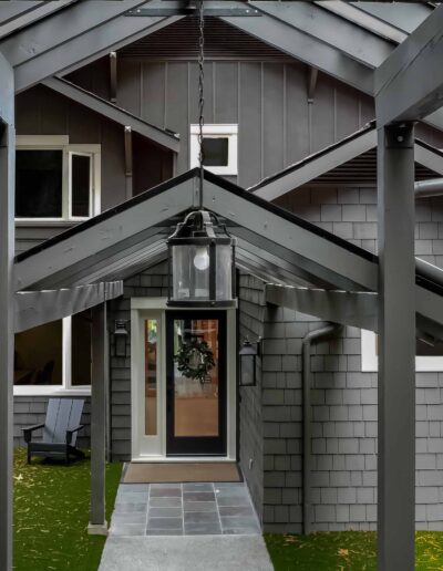 A walkway with a peaked metal roof leads to the front door of a gray house with shingle siding and white trim, surrounded by greenery.