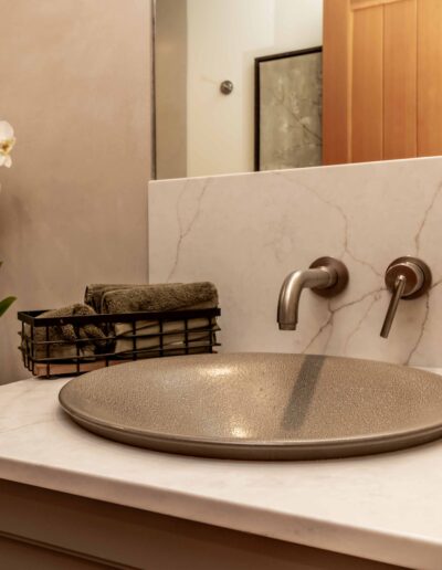 Bathroom vanity with a round gray sink, wall-mounted faucet, soap dispenser, folded towels in a wire basket, and a potted orchid on the counter.