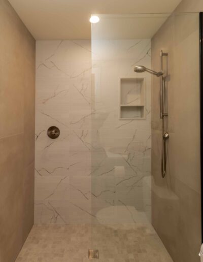 Modern walk-in shower with beige tiled walls, a glass partition, built-in shelves, and a wall-mounted adjustable showerhead.