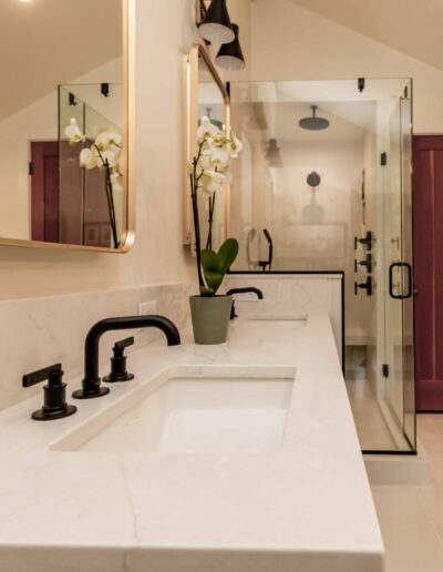 Modern bathroom with a double sink vanity, black fixtures, a potted orchid, large mirrors, and a glass-enclosed shower with a red wooden door in the background.