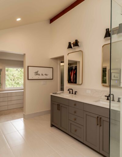 Modern bathroom with double sinks, large mirrors, glass shower, neutral tones, and a view into a walk-in closet with a dresser and framed photos on the wall.