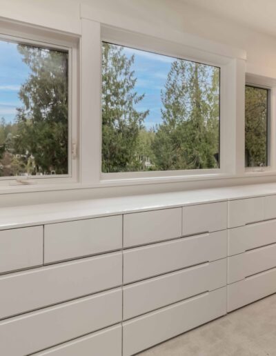 A set of white dressers with multiple drawers sits below three large windows overlooking trees in a bright, modern room.