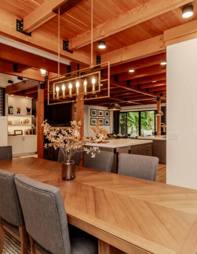 Modern dining room with a wooden table, gray upholstered chairs, overhead chandelier, exposed wooden beams, and an open view into the kitchen and staircase.