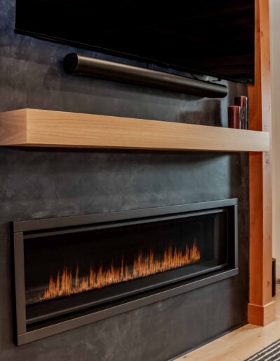 Modern electric fireplace with a linear flame below a light wood mantel, black wall, and a mounted TV above. A striped rug is partially visible on the floor.