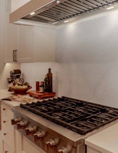 Modern kitchen with a stainless steel gas stove, white cabinets, marble backsplash, and countertop items including a mixer, fruit bowl, and bottles of oil and vinegar.