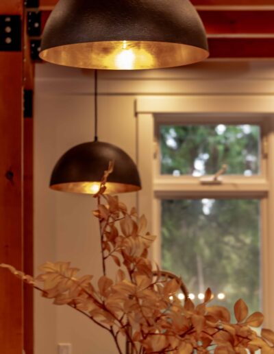 Two black dome pendant lights hang from the ceiling above a vase with dried beige leaves, with a window and greenery in the background.