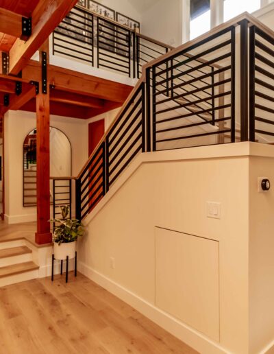 Modern interior with wood floors and exposed beams, metal stair railing, and natural light from large windows. Plant near the stairs and recessed lighting overhead.
