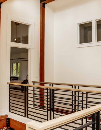 A modern indoor balcony with metal railings, wooden posts, and two windows on white walls, overlooking a lower level.