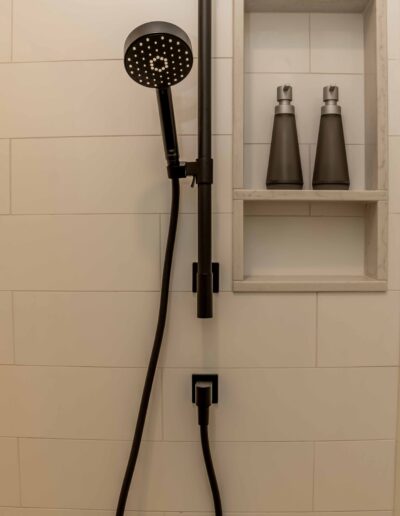 A black handheld showerhead with hose mounted on white tiled wall next to a built-in shelf holding two dark-colored bottles.