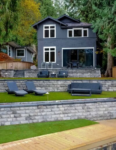 Two-story dark blue house with large windows, tiered stone retaining walls, patio furniture, artificial grass, and surrounded by tall trees and wooden fencing.